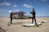Man staat met een peddel in de hand op een subboard op het strand. Andere man legt roeibeweging staand uit.